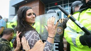 Elle holds her hands open and up in front of a line of police in riot gear. There are people behind her who are also holding their hands up. All hands appear to be white.