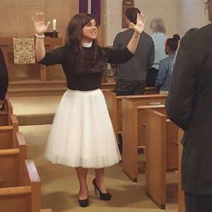 Pastor Elle blesses people during worship. She is a white woman with long brown hair. She is wearing a white tulle skirt and a black clergy shirt with black heels.
