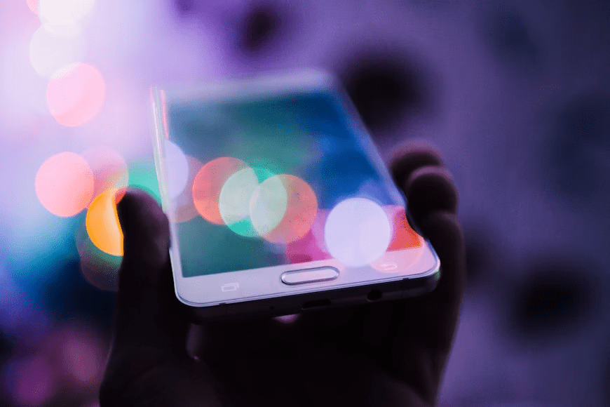 Photo is a stylized image of a person's hand holding a cellphone, with blurry, multicolored lights reflecting around it.